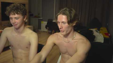 Media: Video of two shirtless young men, one with curly hair and the other with shoulder-length hair, in a dimly lit living room.