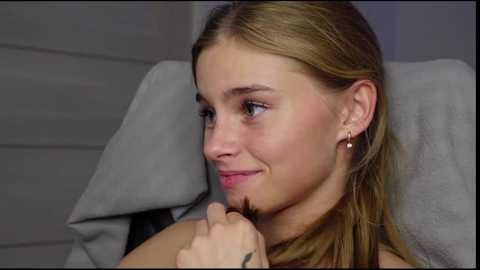 Media: Video of a young, light-skinned woman with blonde hair, wearing a grey top, holding a black hairbrush near her chin, smiling, sitting on a grey cushioned chair.