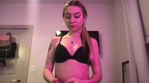 Media: Video of a young Caucasian woman with light skin and long brown hair, wearing a black bra, standing in a dimly lit room with a TV on the wall.