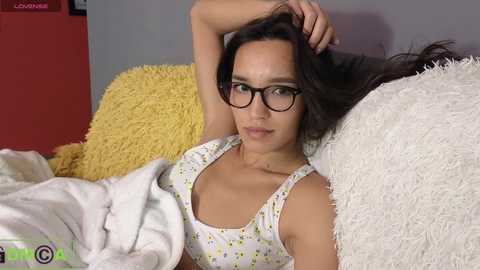 Media: A video of a young woman with light brown skin, wearing glasses, a floral top, and black bra, lying on a bed with yellow and white pillows, in a cozy bedroom.