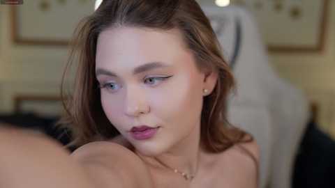 Media: A close-up video of a young, fair-skinned woman with light brown hair, wearing eyeliner and subtle makeup, gazing intently. Background shows a blurred, elegantly furnished room with a cream-colored chair.