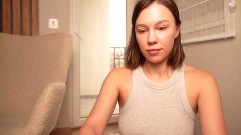 Media: Video of a fair-skinned woman with shoulder-length brown hair, wearing a sleeveless, light gray tank top, sitting on a beige couch in a softly lit room with beige walls and a window with white blinds.