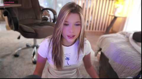 Media: Video of a young, light-skinned woman with long, straight, blonde hair, wearing a white T-shirt with palm tree design, kneeling on a carpeted floor in a modern living room.