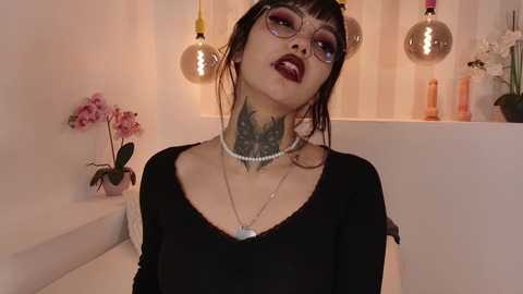 Media: Video of a pale-skinned woman with dark hair and bangs, wearing a black top, large glasses, and a pearl necklace, sporting a detailed tattoo on her neck, in a modern bathroom with pink orchids and hanging pendant lights.