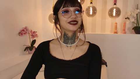 Media: Video of a young woman with pale skin, dark hair, and large glasses, wearing a black top and pearl necklace, tattoos on her neck, in a modern bathroom with pink orchids, glass pendants, and white walls.