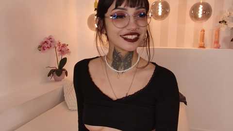 Media: Video of a smiling, pale-skinned woman with black hair, glasses, black top, pearl necklace, and floral tattoo on neck, standing in a modern bathroom with white walls, pink orchid, and metallic spheres.