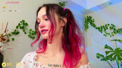 Media: Video of a young woman with long, vibrant pink and black hair, wearing a floral off-shoulder top, standing in a green, plant-filled room.