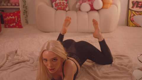 Media: Video of a blonde woman with long hair in a black bodysuit, lying on her stomach with legs bent and feet in the air, in a cozy, warmly lit living room with white furniture and colorful pillows.