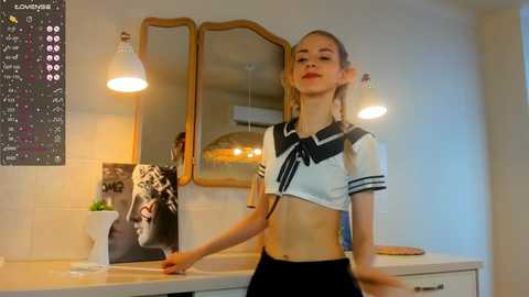 Media: Video of a slender, blonde woman in a cropped sailor top, standing in a modern kitchen with white countertops and hanging pendant lights.