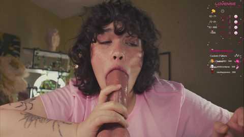 Media: Video of a young, curvy woman with curly dark hair, wearing a pink t-shirt, performing oral sex on a large erect penis, in a dimly-lit room with a live streaming overlay.