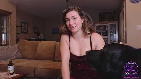 Media: Video of a smiling Caucasian woman with shoulder-length brown hair, wearing a low-cut maroon top, seated on a brown couch, with a black dog sitting beside her.