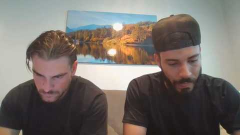 Media: Video of two men with facial hair, wearing black t-shirts, seated indoors. Background features a large, colorful landscape painting of a serene lake and autumn trees.