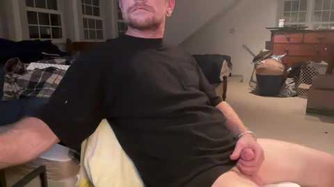 Media: Video of a bearded, white man in a black T-shirt, sitting with legs apart, displaying his circumcised penis in a cluttered, dimly lit room with a bed, dresser, and model airplane.