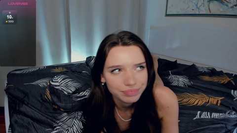 Media: A video of a young woman with long brown hair, wearing a black top, lying on a bed with black and white leaf-patterned sheets in a dimly lit room.