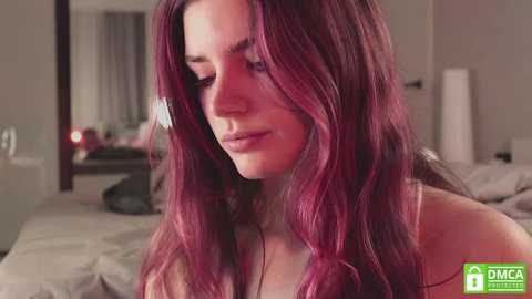 Media: Video of a fair-skinned woman with long, wavy, deep red hair, wearing a white tank top, gazing thoughtfully in a dimly lit bedroom with a bed and a mirror.