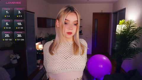 Media: Video of a blonde woman in a white knit top, standing in a modern living room with purple lighting, green plants, and a purple balloon.