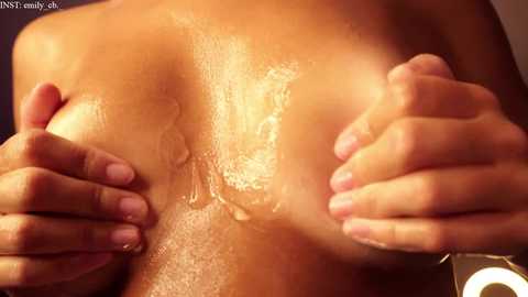 Media: Close-up video of a woman with light brown skin, glistening with oil, using both hands to squeeze her large, firm breasts. The background is dark, possibly indoors.