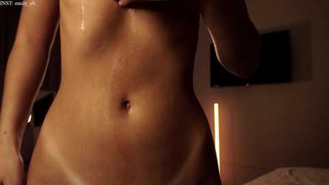 Media: A video of a nude woman with light skin and a toned physique, standing in a dimly lit room. Her body glistens with sweat, highlighting her curves and abdomen. A TV is mounted on the wall behind her.