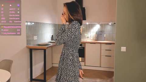 Media: Video of a woman in a black floral dress standing in a modern kitchen, speaking on a phone, with a digital display showing chat messages in the background.