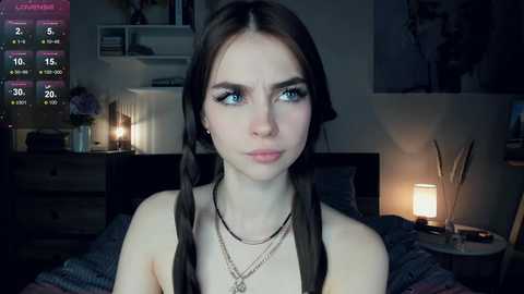 Media: Video of a young woman with pale skin, blue eyes, and dark brown hair in twin braids, wearing a black top and multiple necklaces, indoors with dim lighting, a bed, and a lit lamp in the background.