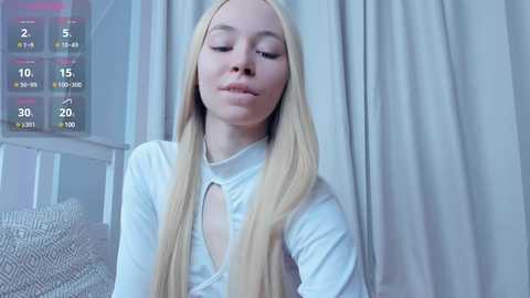 Media: Video of a young Caucasian woman with long, straight blonde hair, wearing a white top, standing in a light-filled room with a white curtain backdrop.