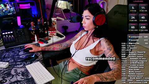 Media: Video of a tattooed woman with medium breasts wearing headphones, seated at a gaming setup in a dimly lit room, streaming on Twitch with various chat messages displayed.