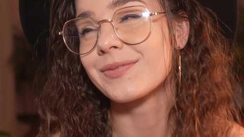 Media: Video of a smiling young woman with curly brown hair, wearing round glasses and long gold earrings, set against a softly blurred indoor background.