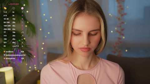 Media: A video of a blonde woman with fair skin and a slender physique, wearing a light pink top, sitting indoors with a serene expression.