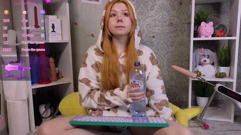 Media: Video of a fair-skinned, red-haired woman in a bear-themed pajama, sitting cross-legged on a yellow chair, holding a water bottle, in a cluttered room with patterned wallpaper and shelves.