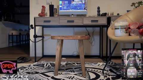 Media: Video of a messy modern living room with a wooden desk, a computer monitor, and a pink neon sign. A black chair with a white cushion is nearby, and a wine bottle is on the floor.