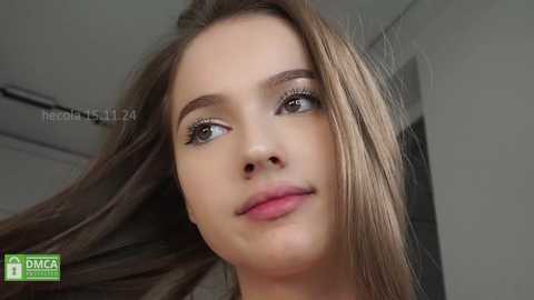 Media: Video of a young Caucasian woman with light skin, brown hair, and green eyes, looking slightly to the right. She has a neutral expression and is indoors with a blurred background.