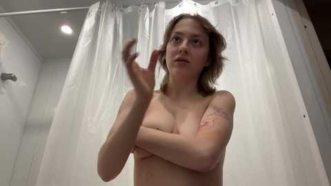 Media: Video of a young, slender, light-skinned woman with shoulder-length brown hair, topless, standing in a shower with white curtain. She has a tattoo on her upper arm and is adjusting her hair.