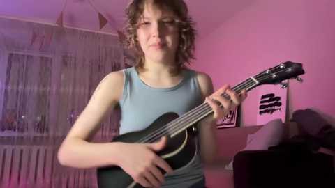 Media: Video of a young Caucasian boy with curly brown hair, playing an electric guitar in a dimly lit room with pink lighting. He wears a blue tank top.