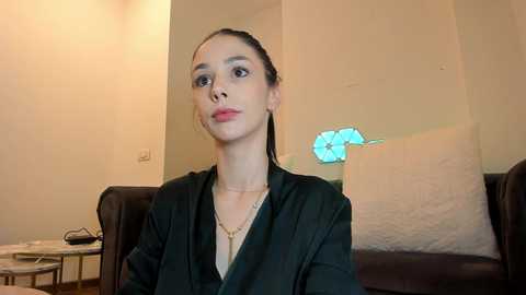 Media: Video of a slender woman with light skin, dark hair pulled back, wearing a black blazer over a beige top, seated indoors. Background features beige walls and a blue flowered cushion.