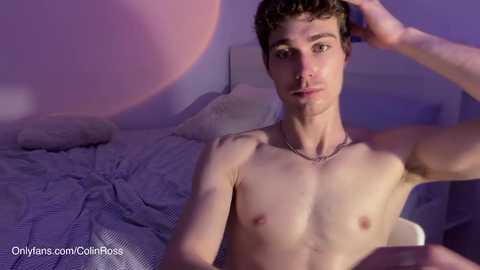 Media: Video of a shirtless, slim, young white man with curly hair, sitting on a bed with a gray bedspread, looking at the camera.