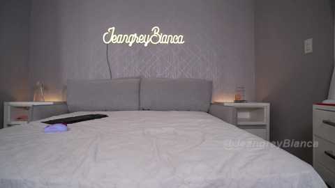 Media: Video of a modern bedroom with a grey upholstered bed, white nightstands, a \"Jamprey Bianca\" neon sign, and a remote control on the bed.