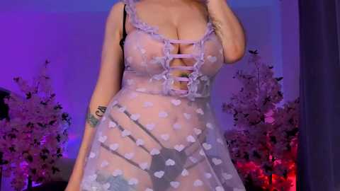 Media: Video of a woman in sheer, white polka-dot lingerie, exposing cleavage, with a tattoo on her left arm. Background features pink cherry blossom trees and purple lighting, creating a romantic atmosphere.