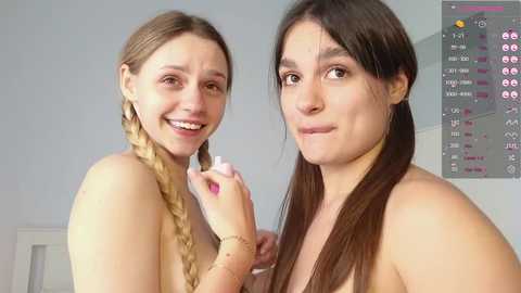 Media: Video of two smiling women with fair skin and long hair, one with a braided ponytail, topless, standing in a brightly lit room with a calendar in the background.