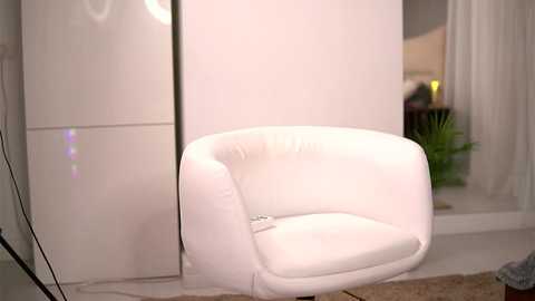 Media: Video of a modern white leather armchair in a minimalist room with cream walls, a white wardrobe, and a potted plant on the floor.