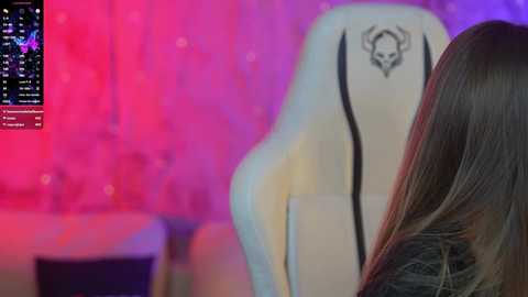 Media: Video of a woman with long brown hair in a gaming chair with a skull logo, against a vividly lit pink and purple backdrop.