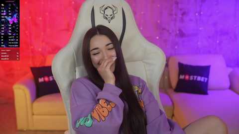 Media: Video of a young woman with long black hair, wearing a purple hoodie, seated in a white gaming chair, illuminated by colorful LED lights, with a video game screen on the left.