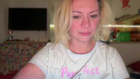 Media: Video of a blonde Caucasian woman with shoulder-length hair, wearing a gray T-shirt with pink writing. She stands in a cozy living room with colorful, patterned curtains, a green plant, and a flat-screen TV on the wall.