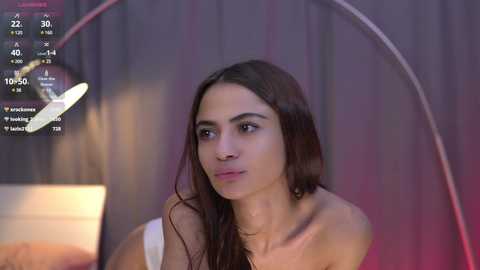 Media: Video of a young, dark-haired woman with light skin, wearing a strapless top, sitting in a dimly lit room with a modern, curved lamp and a digital clock showing 10:05.