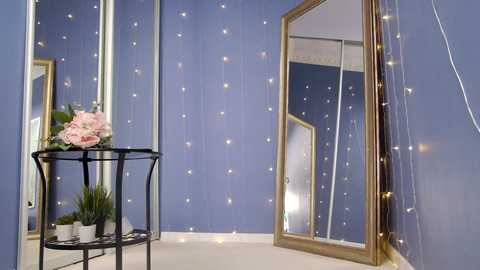 Media: A video of a serene, blue-walled room with fairy lights, a black wrought-iron plant stand, and a large gold-framed mirror.