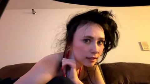 Media: Video of a woman with black hair, red lipstick, and a red necklace, lying on a brown couch in a dimly lit room.