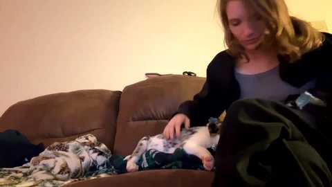 Media: Video of a woman with shoulder-length blonde hair, wearing a gray shirt, sitting on a brown leather couch, comforting a white and gray kitten in her lap.