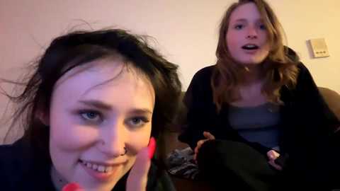 Media: A video of two young women with pale skin and light brown hair, one smiling, the other talking animatedly. They are indoors, in a casual setting with a beige wall and a light switch visible.