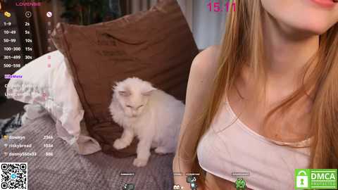 Media: Video of a light-skinned woman with long, straight blonde hair, wearing a white tank top, sitting on a bed with a fluffy white cat. Background includes a brown pillow and a grey carpet.