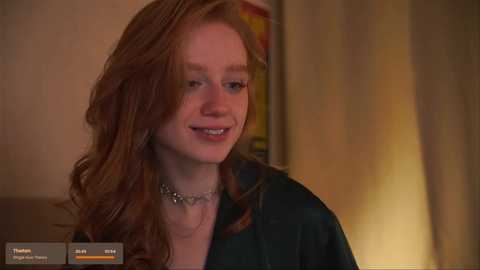 Media: Video of a smiling, fair-skinned young woman with long, wavy red hair, wearing a dark green jacket and silver necklace, indoors with a blurred background.