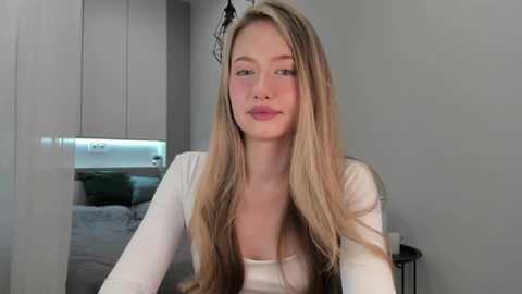 Media: Video of a young Caucasian woman with long, wavy blonde hair and fair skin, wearing a white top, standing in a modern kitchen with light gray cabinets and a stainless steel oven.